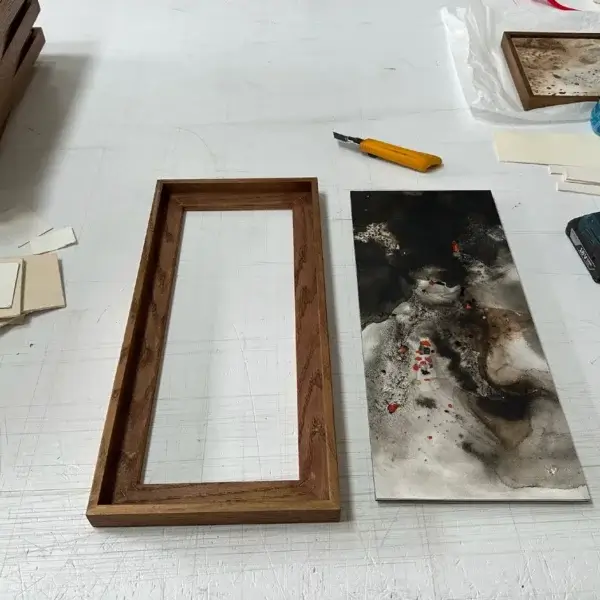 Caisse américaine en chêne prête pour l'encadrement d'une œuvre de Léa Aquilina marouflée sur aluminium, avec l'œuvre et les outils d'atelier. En arrière-plan, œuvres déjà encadrées, atelier Atelier Annel."